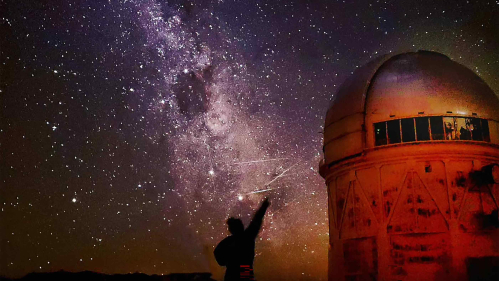 Person silhouetted against the sky with stars above and an observatory to the right