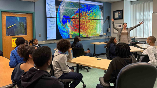 Woman standing in front of screen with world map speaking to a group of students