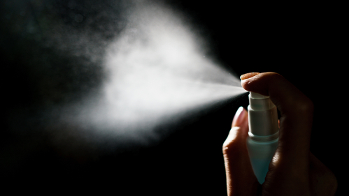 Hand at the right shown pushing the lever of a spray bottle. Spray is emitted.