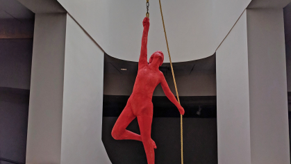 Life-sized sculpture of an acrobat painted red, hanging from ceiling by a yellow rope near a skylight 