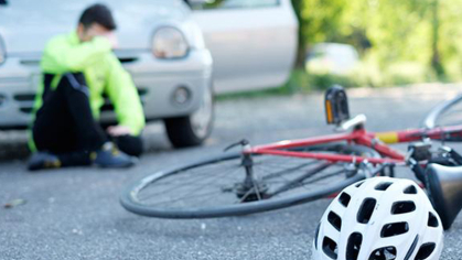 Bike on the ground with a helmet and a person in the background sitting in front of a car after being hit