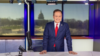 Mike Emanuel standing at the news desk wearing a blue suit and red tie