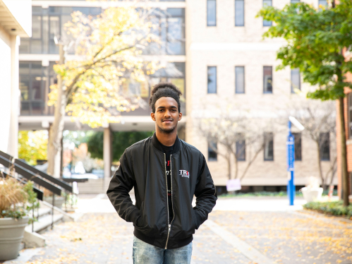 Wandys Aquino standing on the Rutgers-Camden campus wearing a black jacket with the TRIO logo