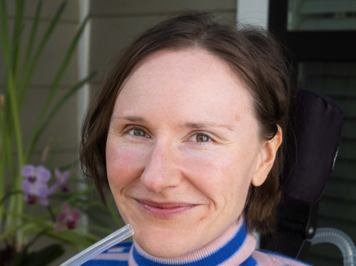 Park McArthur, a white person with short brown hair, smiles while wearing a pink and blue striped sweater. Purple flowers and McArthur's mouthpiece ventilator and powerchair headrest are also visible.