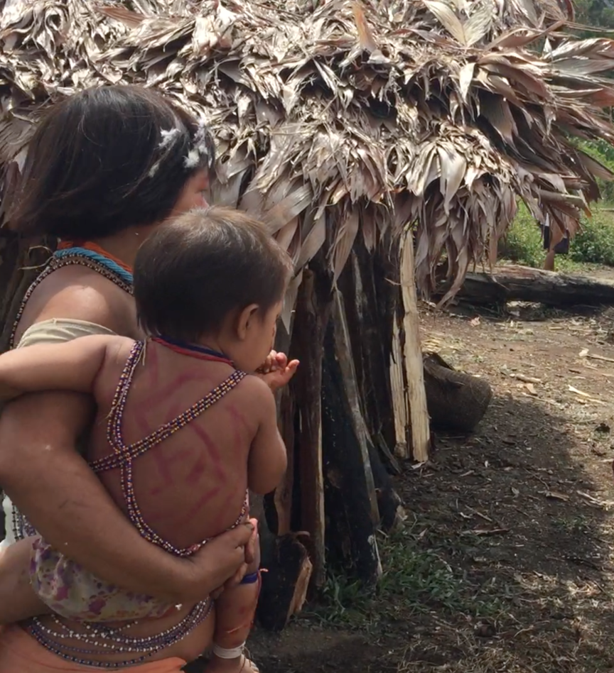 A young adult and toddler stand near a small hut in a Venezuelan rainforest village.