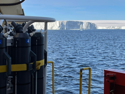 Sampling rosette with gray sampling bottles at left, the ship's rail at lower right, and the face of the ice shelf in the background.