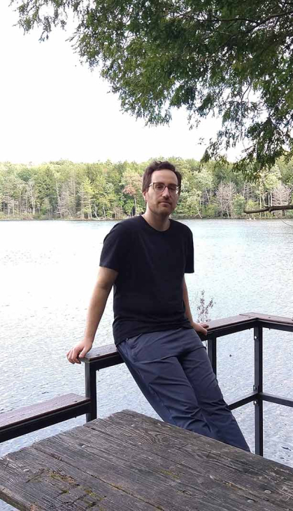 man in dark T-shirt and blue pants leaning against railing in front of a lake.