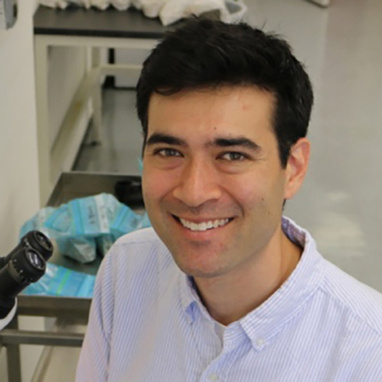 man with light blue oxford button shirt with top button unbuttoned with microscope in background