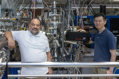 Two men stand in front of a vast network of steel pipes and machines.