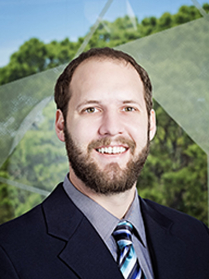 Man with beard, gray shirt, striped tie, and black jacket with trees in the background.