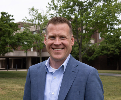 Man in blue jacket stands on campus with grass and trees behind him