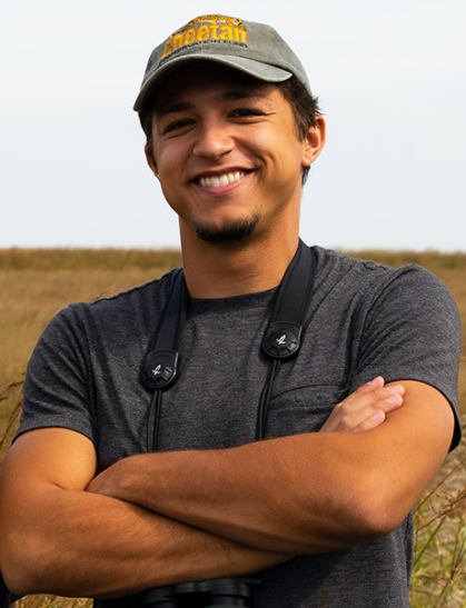 Man wearing baseball cap with arms crossed smiles at camera