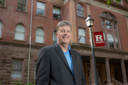 Bill Welsh standing in front of Winants Hall with Rutgers flag in the background