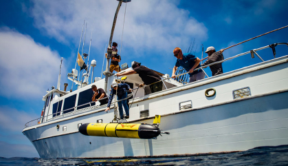 The Sentinel Redwing glider, a yellow submersible research robot is hoisted from a ship to bring it into the ocean.